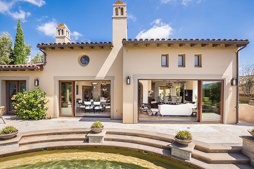 AG Millworks(sliding) Single-story house with beige stucco exterior, arched windows, and double glass doors opening to a patio with steps leading to a circular pool. The sky is clear and the surroundings include plants.