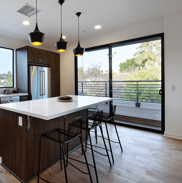 Milgard A modern kitchen with a central island, three bar stools, pendant lights, stainless steel appliances, and large sliding glass doors leading to a balcony with a view of trees.
