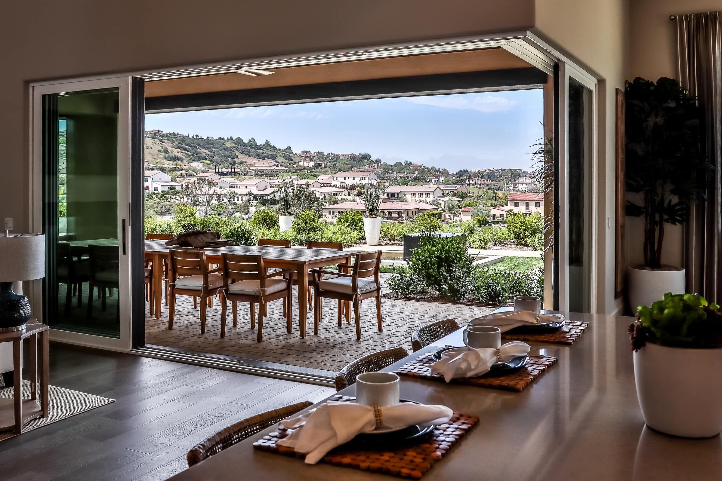 Windor(sliding) Open sliding glass doors reveal an outdoor patio with a dining set overlooking a suburban neighborhood, seen from a modern indoor kitchen with a breakfast bar and place settings.